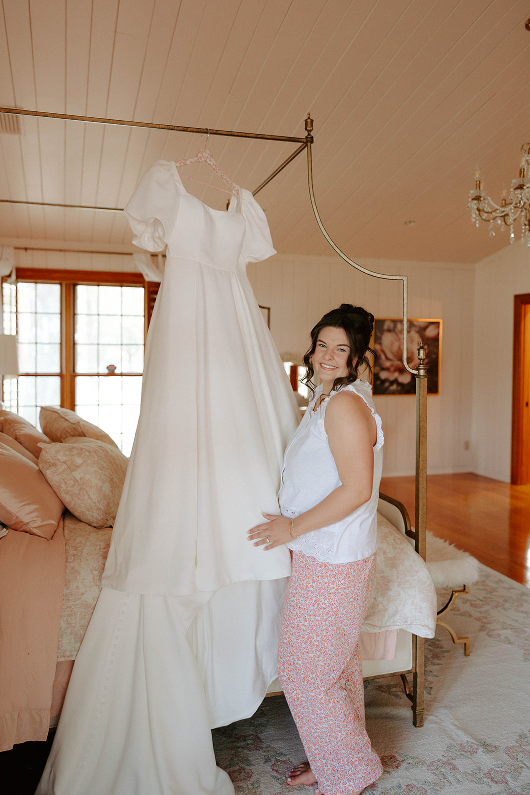 Bride posing with wedding dress hanging from bed canopy